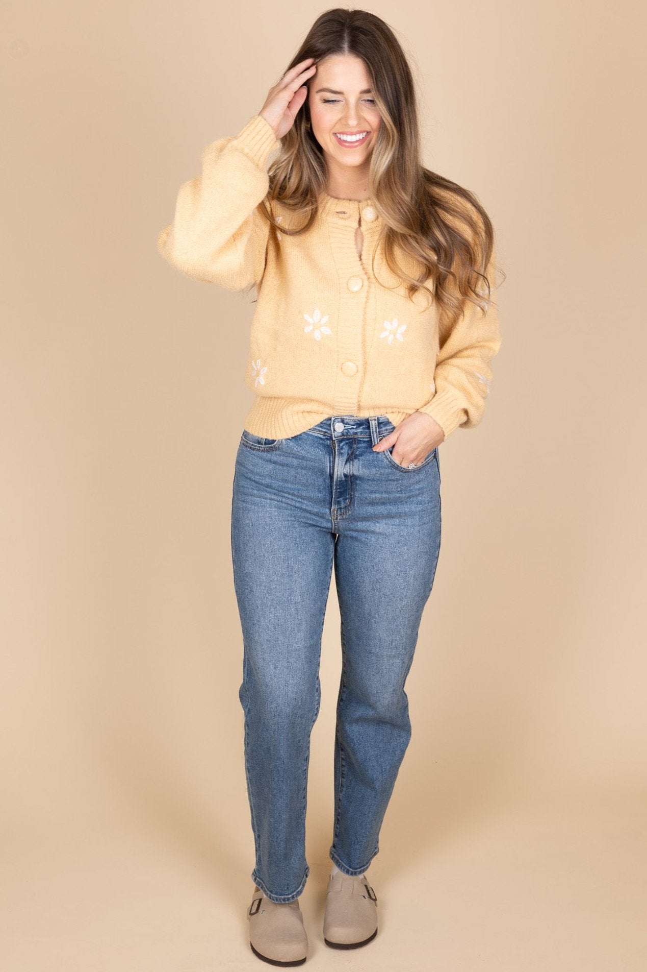 Darlin Daisy Knit Cardigan front view in a light caramel color. Embroidered white daisies are spread evenly throughout sweater. Large button front closure with billowy sleeve. Hits about waist length and is paired with a medium wash jean and clogs.