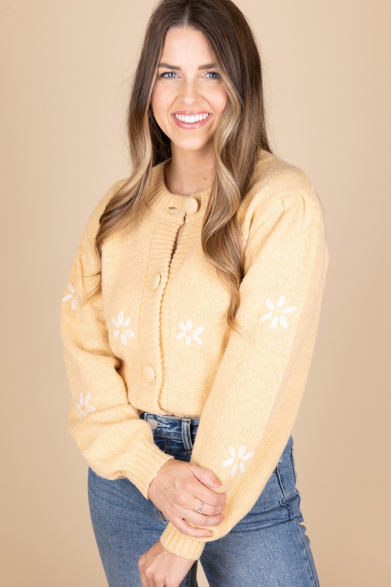 Darlin Daisy Knit Cardigan front view in a light caramel color. Embroidered white daisies are spread evenly throughout sweater. Large button front closure with billowy sleeve. Hits about waist length and is paired with a medium wash jean.