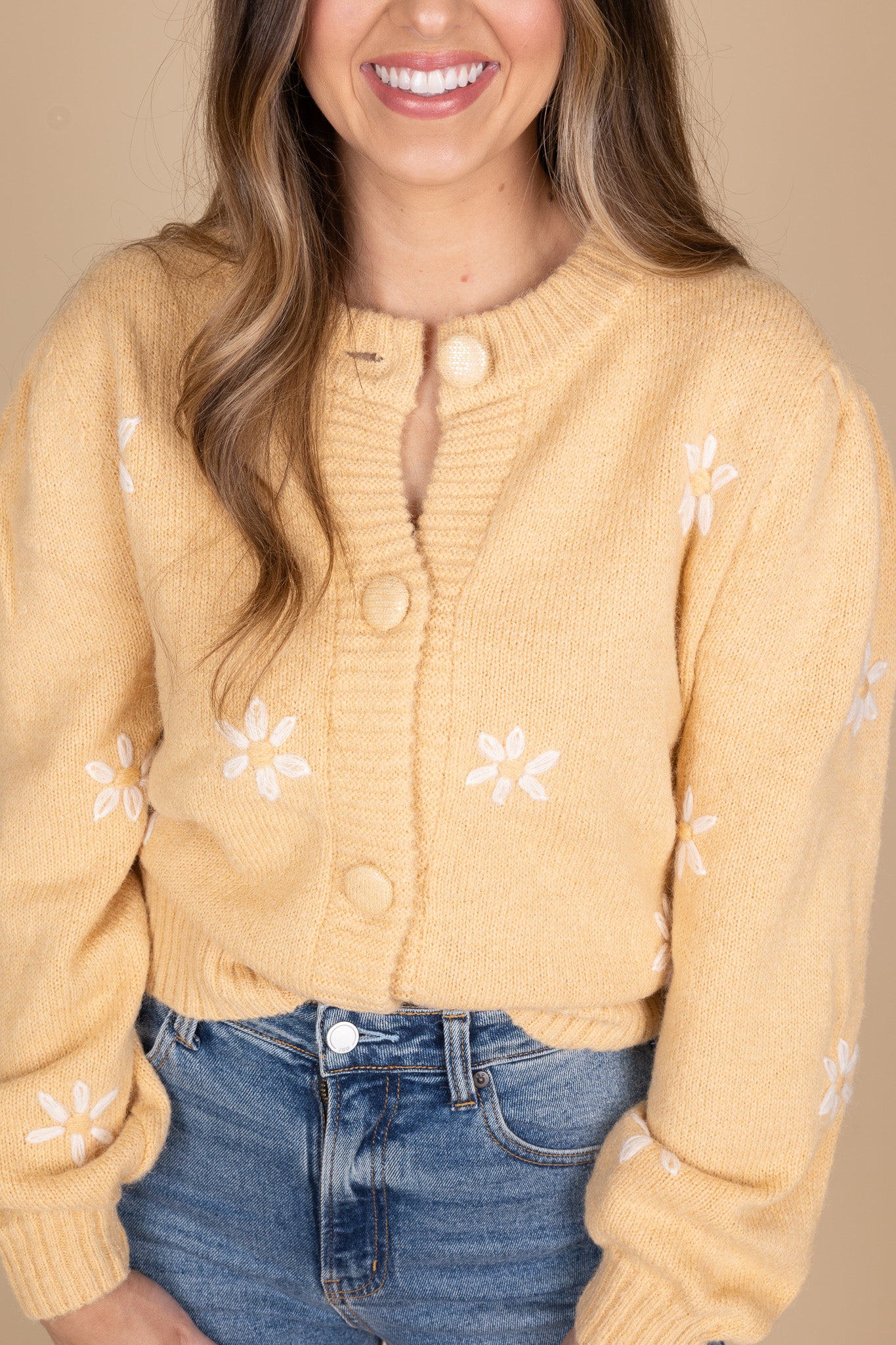 Darlin Daisy Knit Cardigan close up view in a light caramel color. Embroidered white daisies are spread evenly throughout sweater. Large button front closure with billowy sleeve. Hits about waist length and is paired with a medium wash jean.