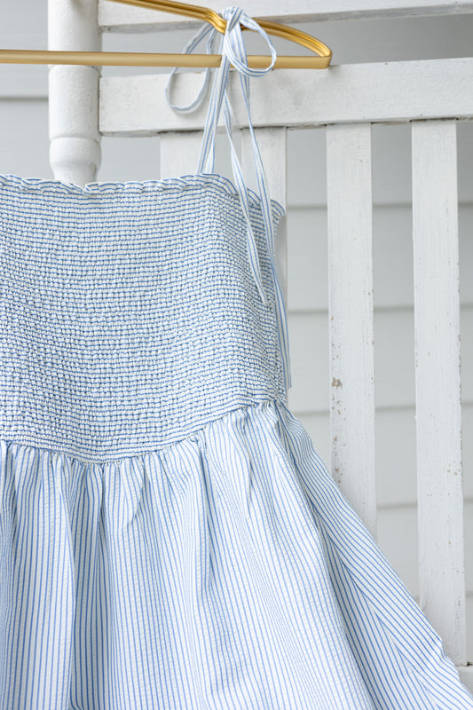 close up Rowan Seersucker Striped Dress in blue and white with bow tie straps, smocked bodice, ruffle hem, and functional pockets