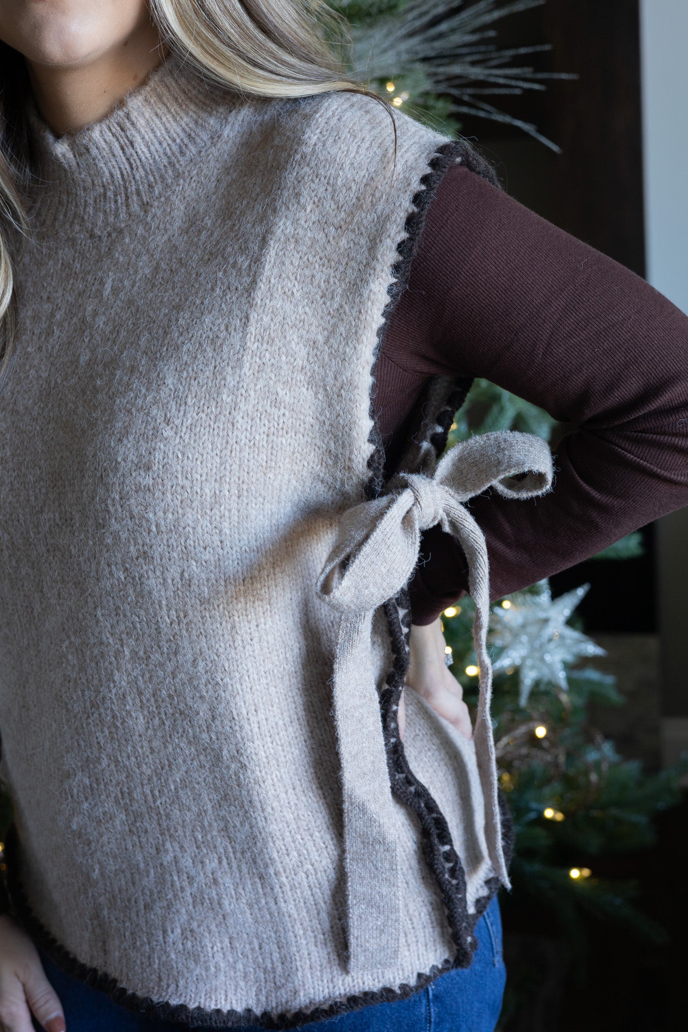 Person wearing a gray knitted vest with a bow detail, standing in front of a decorated Christmas tree.