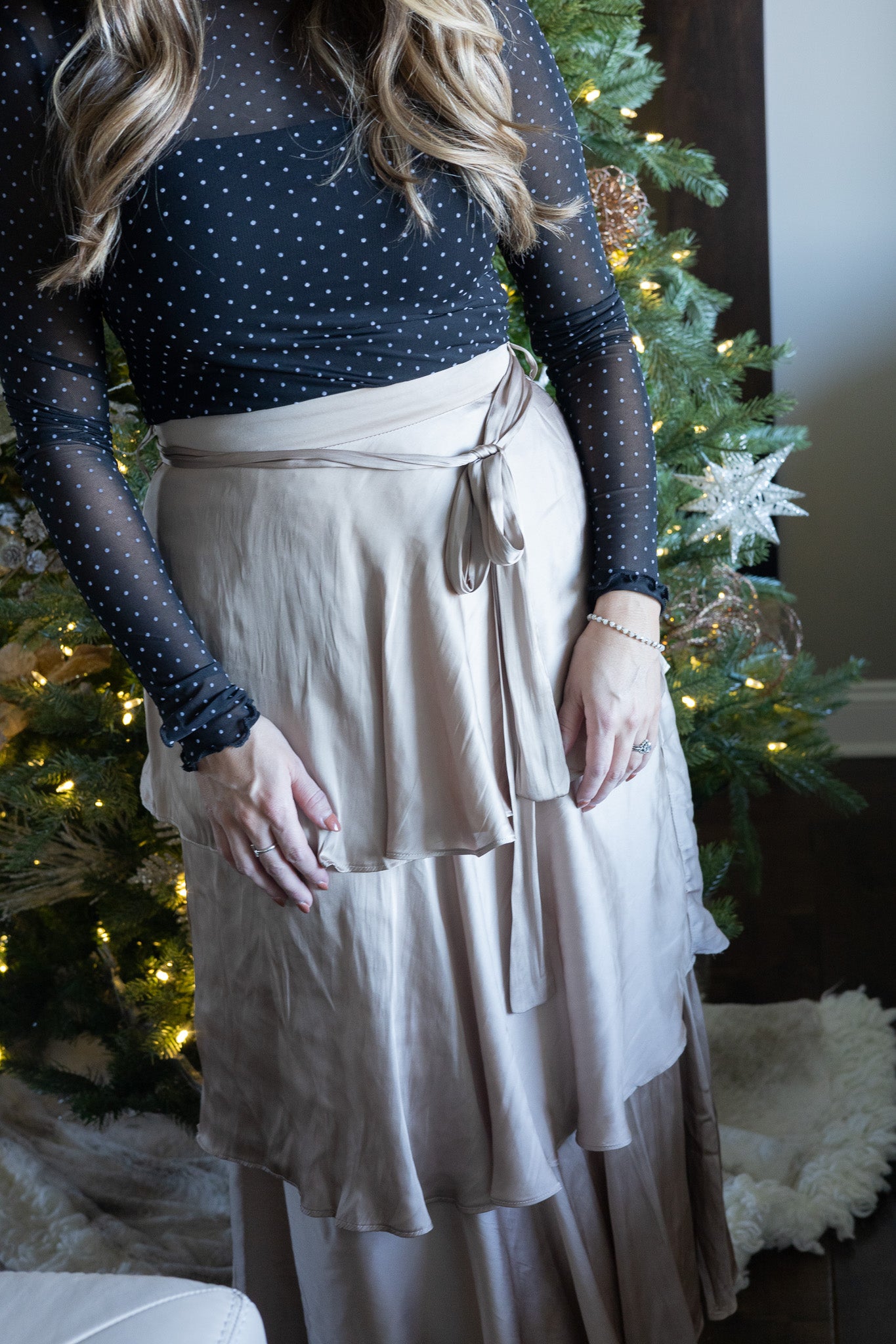 Person wearing a black polka dot top and beige skirt in front of a decorated Christmas tree.
