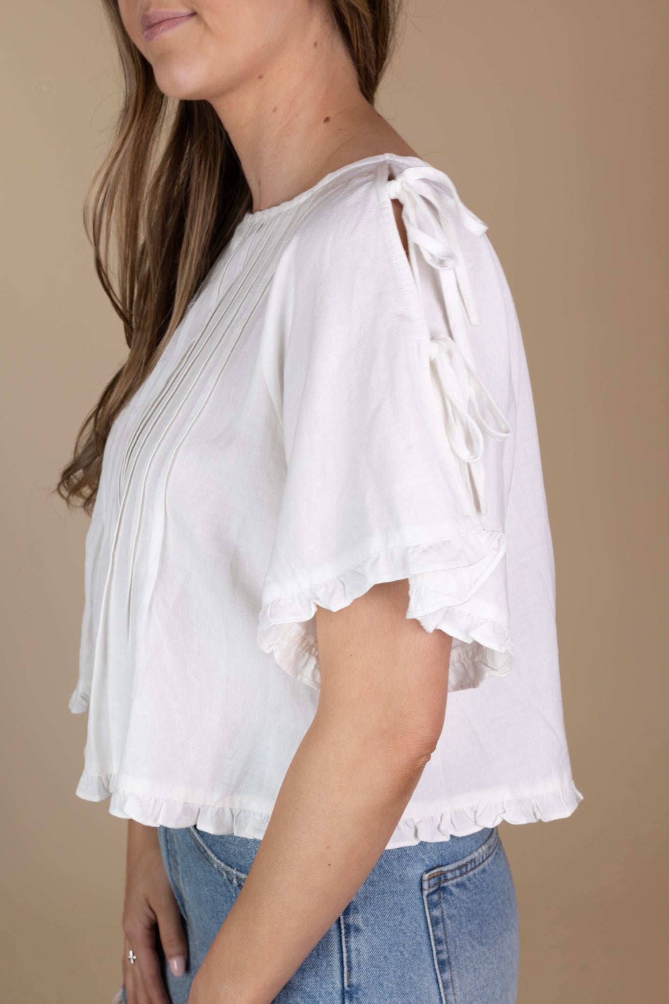 side view of off-white Addie Pleated Blouse with ruffle hem, tie sleeves, and pleated detailing, showcasing a loose bodice and hip-length cut.