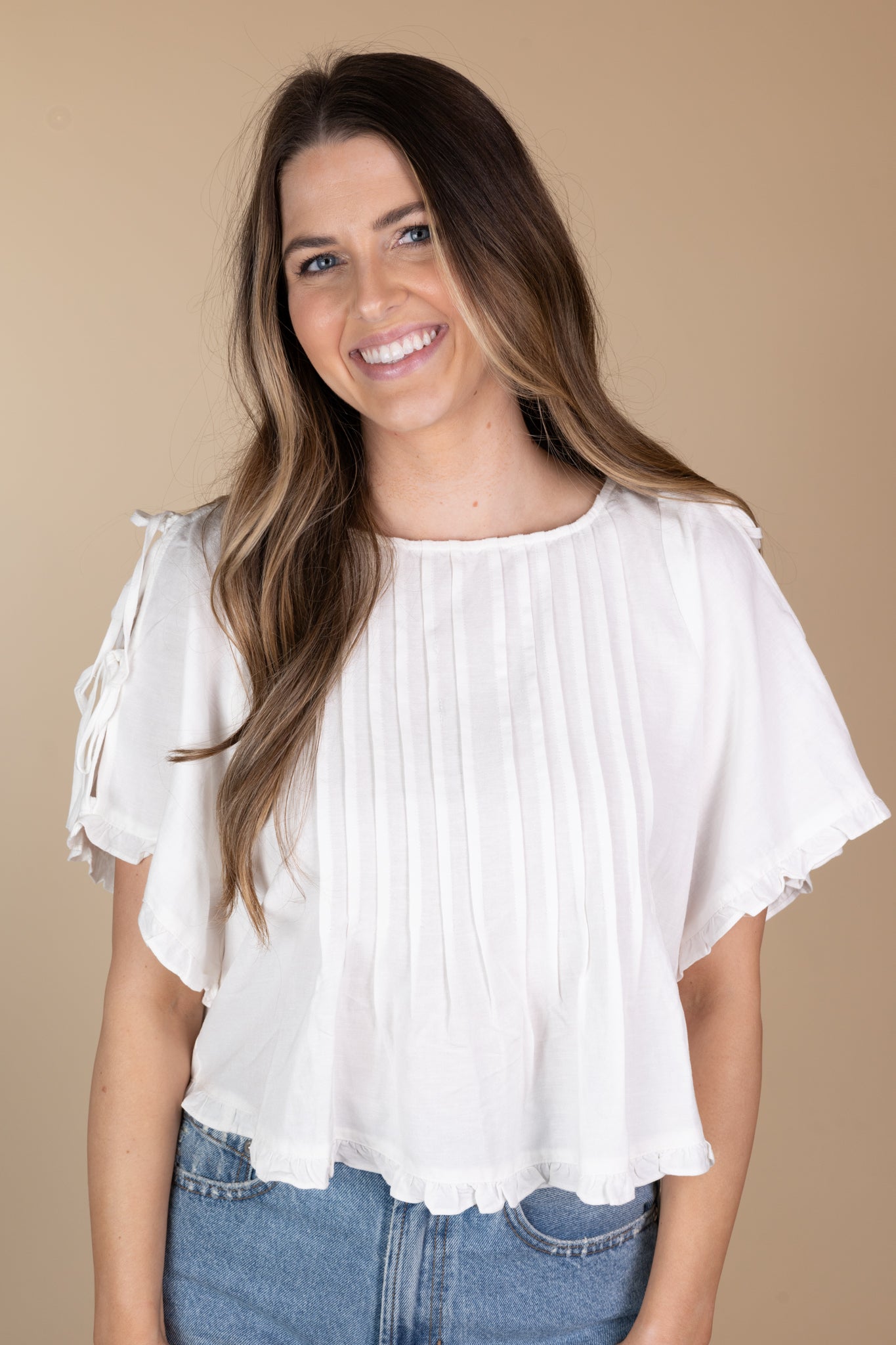 front view of off-white Addie Pleated Blouse with ruffle hem, tie sleeves, and pleated detailing, showcasing a loose bodice and hip-length cut.