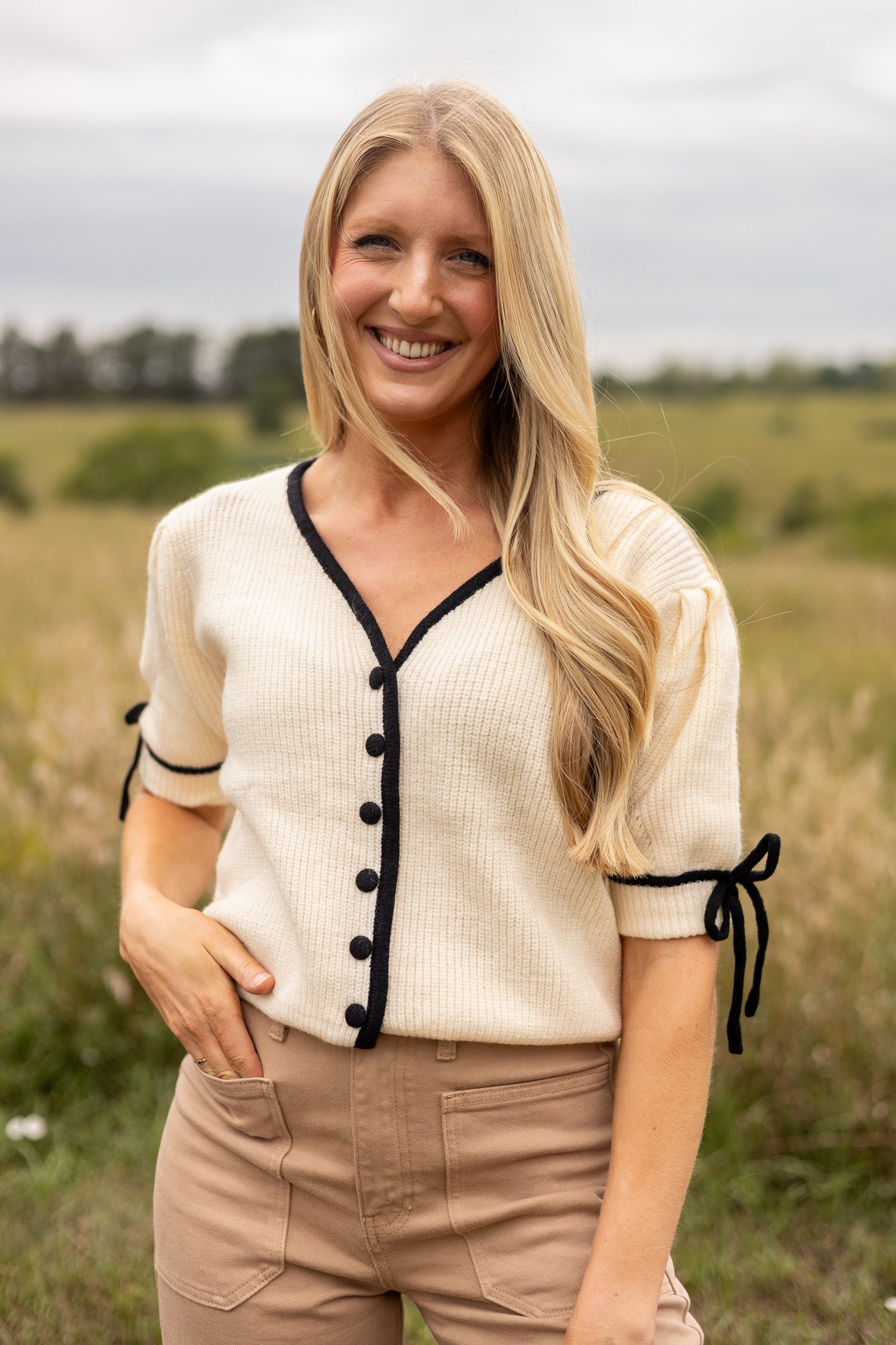 full front view of cream short puff sleeve sweater with front v neck and buttons, has black trim and buttons with black tie bow on sleeves