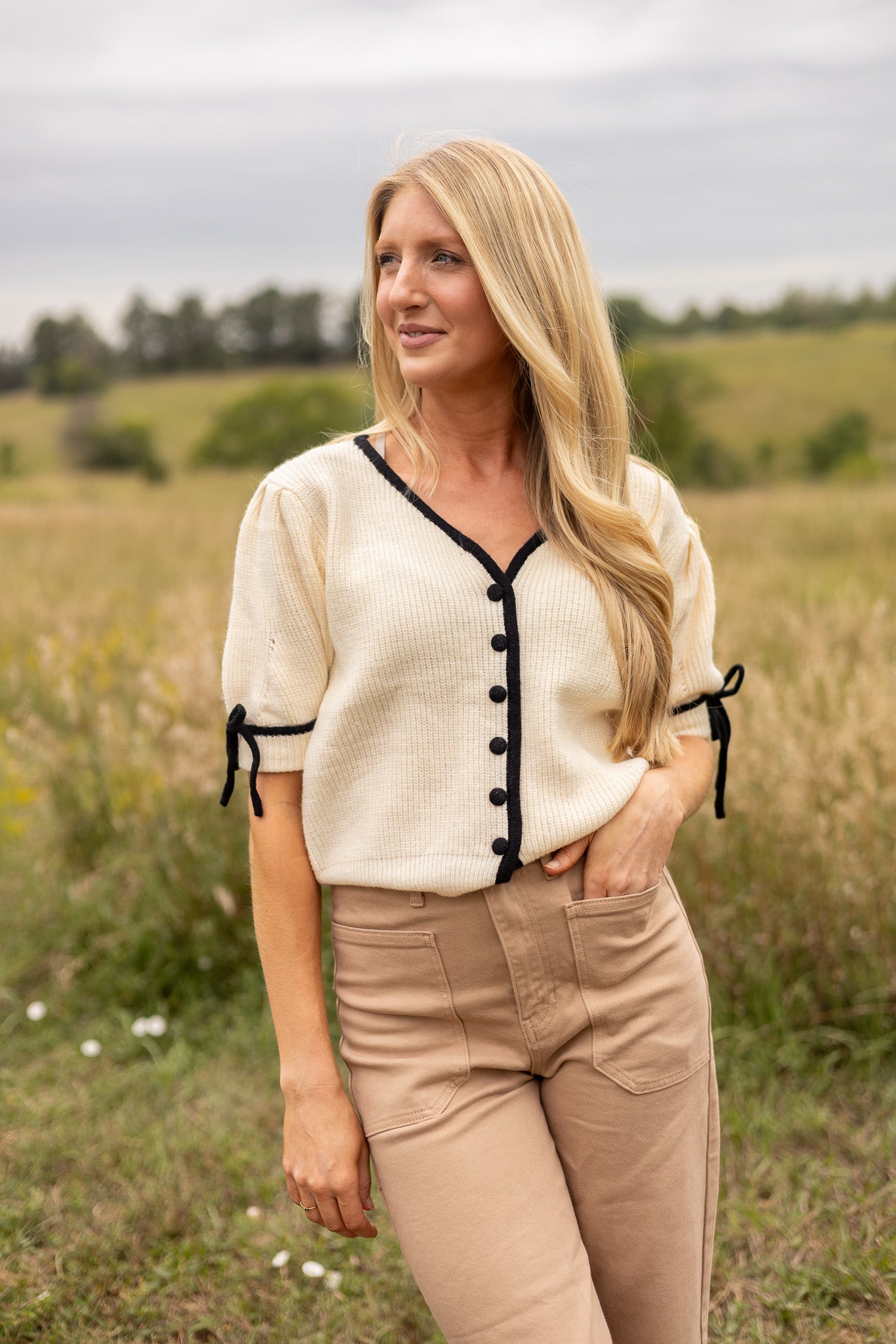 front view of cream short puff sleeve sweater with front v neck and buttons, has black trim and buttons with black tie bow on sleeves, paired with light brown denim