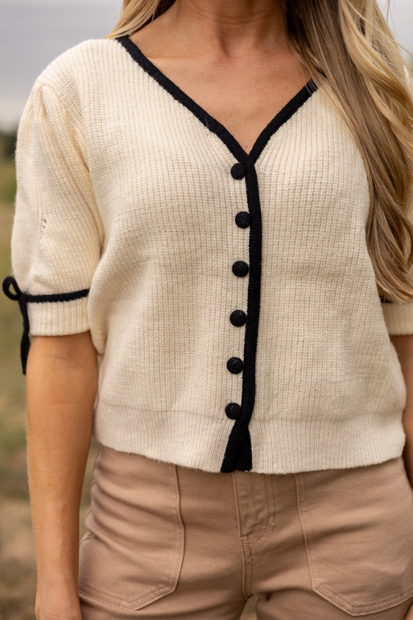 close up view of cream short puff sleeve sweater with front v neck and buttons, has black trim and buttons with black tie bow on sleeves