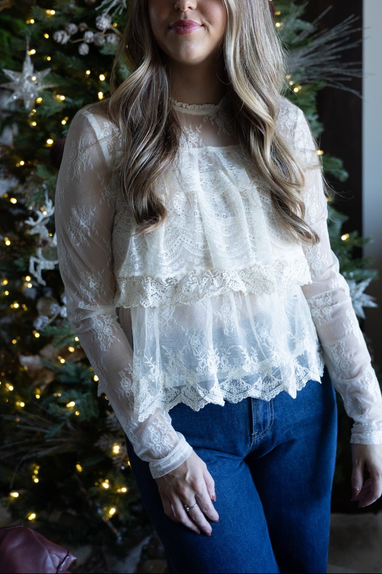 Woman wearing a lace top and blue jeans standing in front of a decorated Christmas tree.