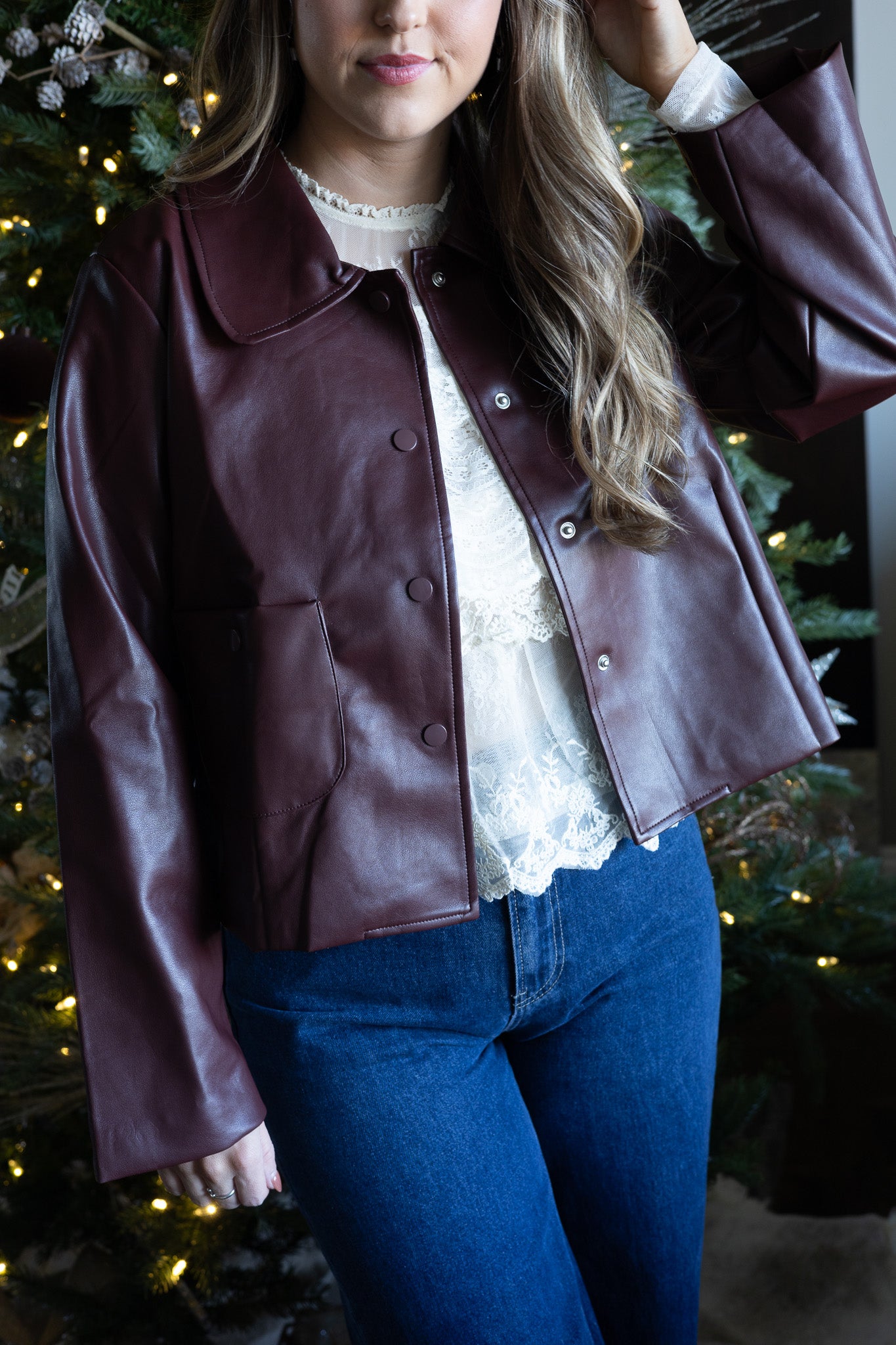 Person wearing a brown leather jacket over a white lace top and blue jeans, standing in front of a decorated Christmas tree.